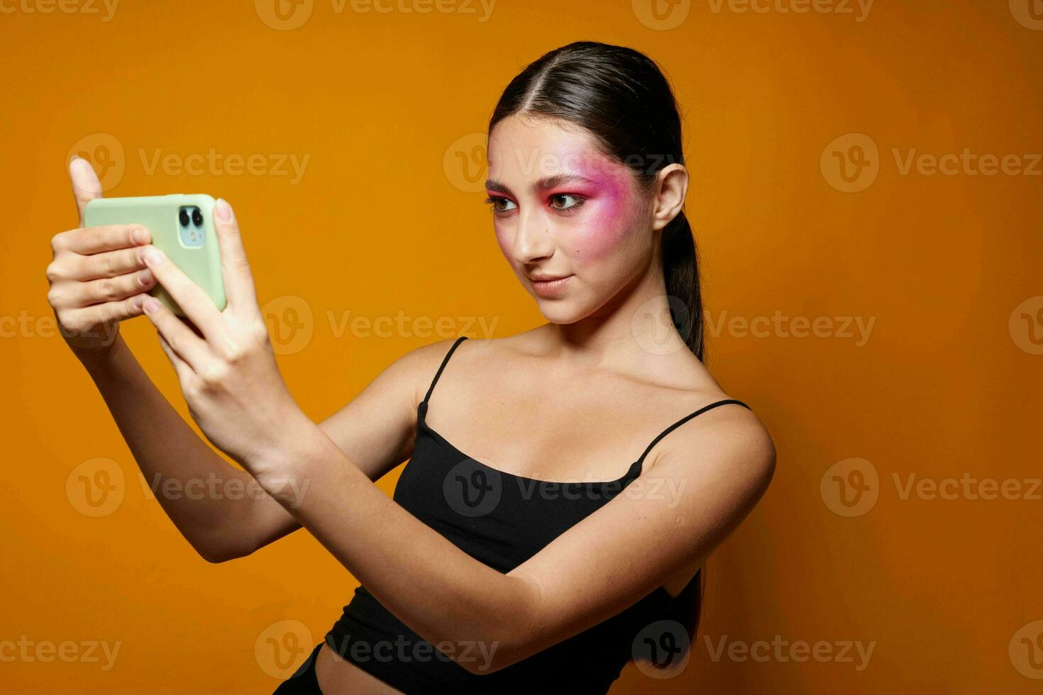 sexy morena mulher brilhante Maquiagem posando Preto jérsei com uma telefone dentro mão amarelo fundo inalterado foto