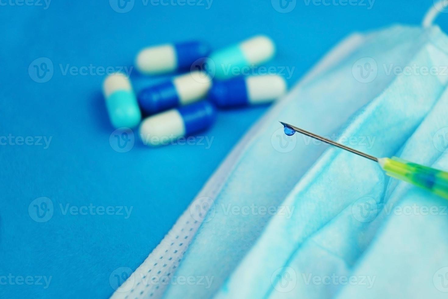 uma macro detalhada de uma seringa por pílulas com uma gota de líquido azul através da agulha em uma máscara médica com fundo azul foto