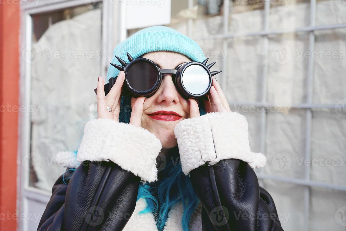 retrato de uma jovem punk ou gótica sorrindo com cabelo de cor azul e usando óculos pretos steampunk e boné de lã azul em uma rua urbana ao ar livre foto