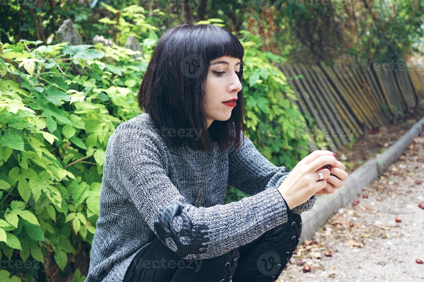 Linda mulher morena caucasiana, sentada no chão em um parque, usando roupas punk ou góticas e cercada por folhas verdes foto