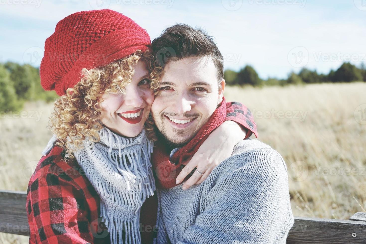 jovem casal de namorados romântico olha para a câmera enquanto sorri e se ama, sentado em um banco de madeira ao ar livre com a natureza como pano de fundo foto