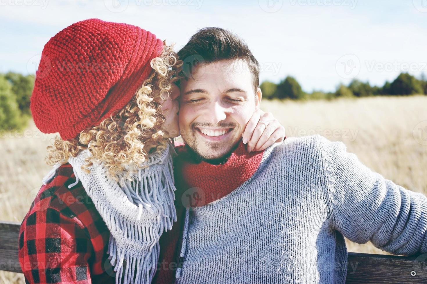 jovem casal romântico de uma linda mulher loira com cabelo encaracolado e um boné de lã vermelha beijando o namorado e um homem bonito ao ar livre foto