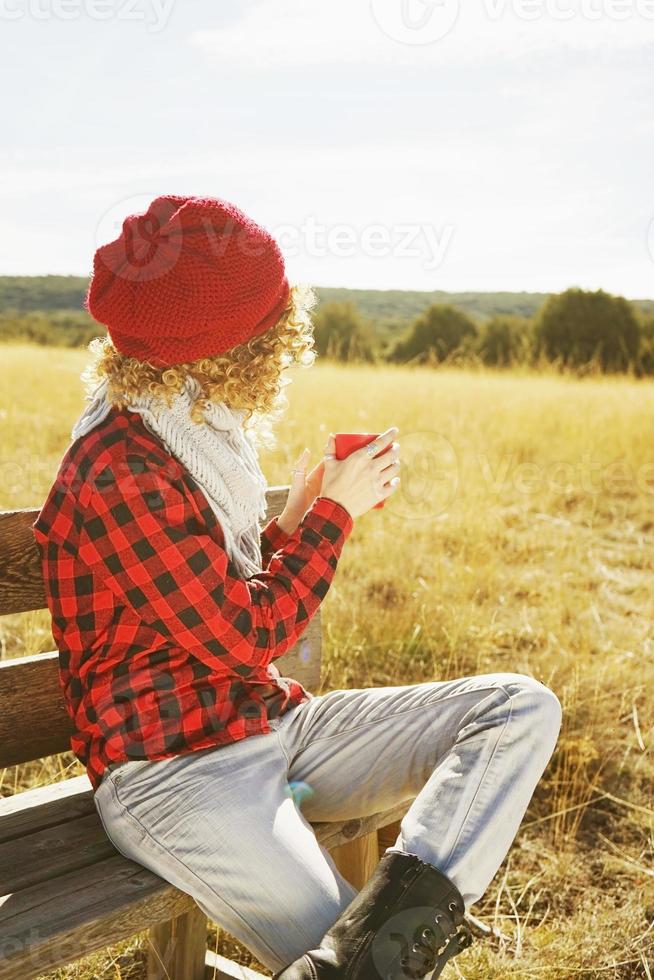 uma jovem mulher por trás com uma camisa xadrez vermelha com um boné de lã e cachecol tomando uma xícara de chá ou café enquanto ela está tomando sol, sentada em um banco de madeira em um campo amarelo com luz de fundo foto