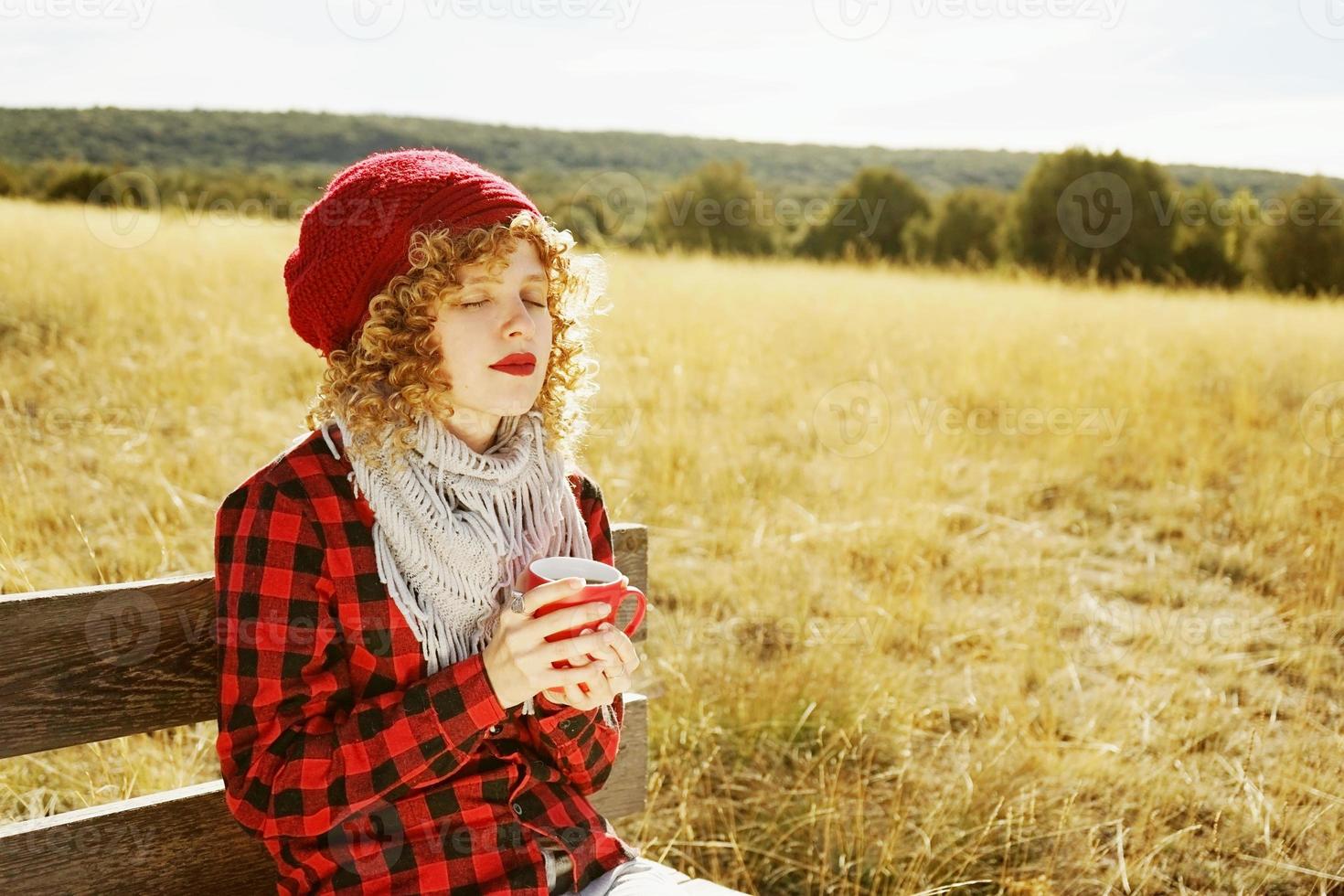 retrato frontal de uma jovem com uma camisa xadrez vermelha com um boné de lã e cachecol tomando uma xícara de chá ou café enquanto ela está se bronzeando, sentada em um banco de madeira em um campo amarelo com luz de fundo do sol de outono foto