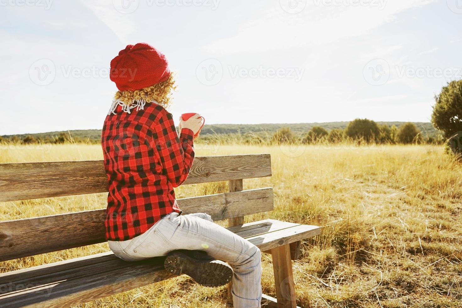 uma jovem mulher por trás com uma camisa xadrez vermelha com um boné de lã e cachecol tomando uma xícara de chá ou café enquanto ela está tomando sol, sentada em um banco de madeira em um campo amarelo com luz de fundo foto