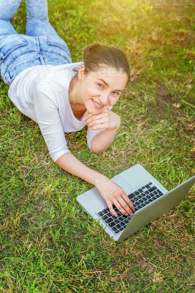 jovem mulher deitado em verde Relva gramado dentro cidade parque trabalhando em computador portátil pc computador. freelance o negócio conceito foto