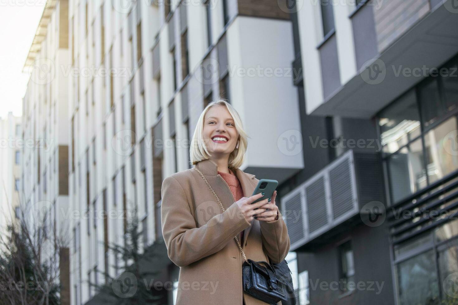 linda lindo jovem mulher com Loiras cabelo Mensagens em a Smartphone às a cidade rua fundo. bonita menina tendo inteligente telefone conversação foto