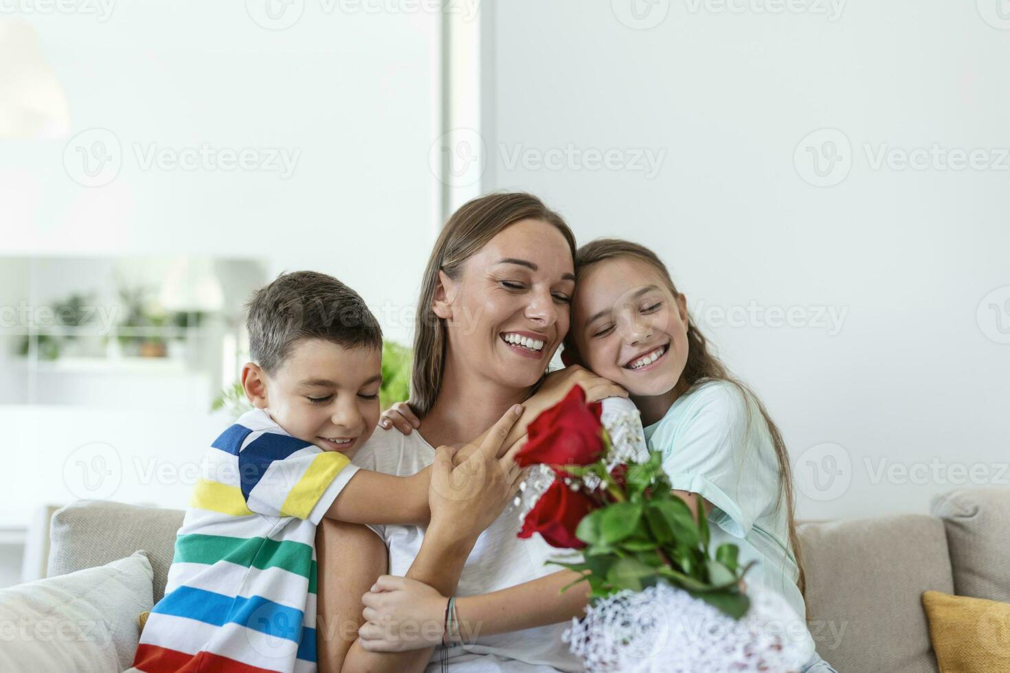 jovem mãe com uma ramalhete do rosas risos, abraçando dela filho, e alegre menina com uma cartão parabeniza mãe durante feriado celebração dentro cozinha às casa foto