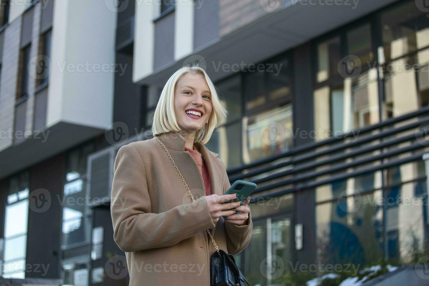 linda lindo jovem mulher com Loiras cabelo Mensagens em a Smartphone às a cidade rua fundo. bonita menina tendo inteligente telefone conversação foto
