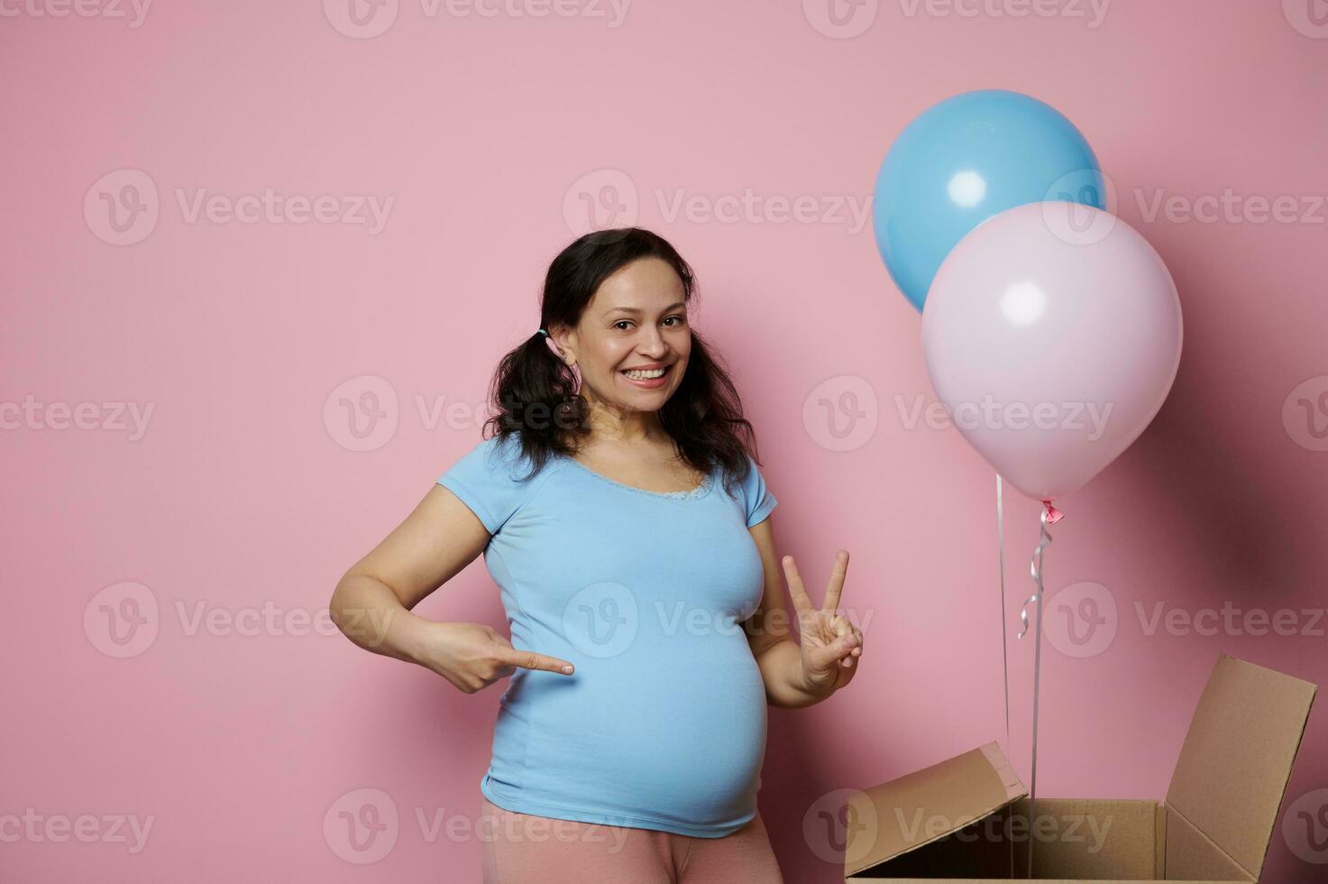 espantado grávida mulher, esperando gêmeos, mostra dois dedos olhando às Câmera, isolado Rosa pano de fundo. gênero revelar festa foto