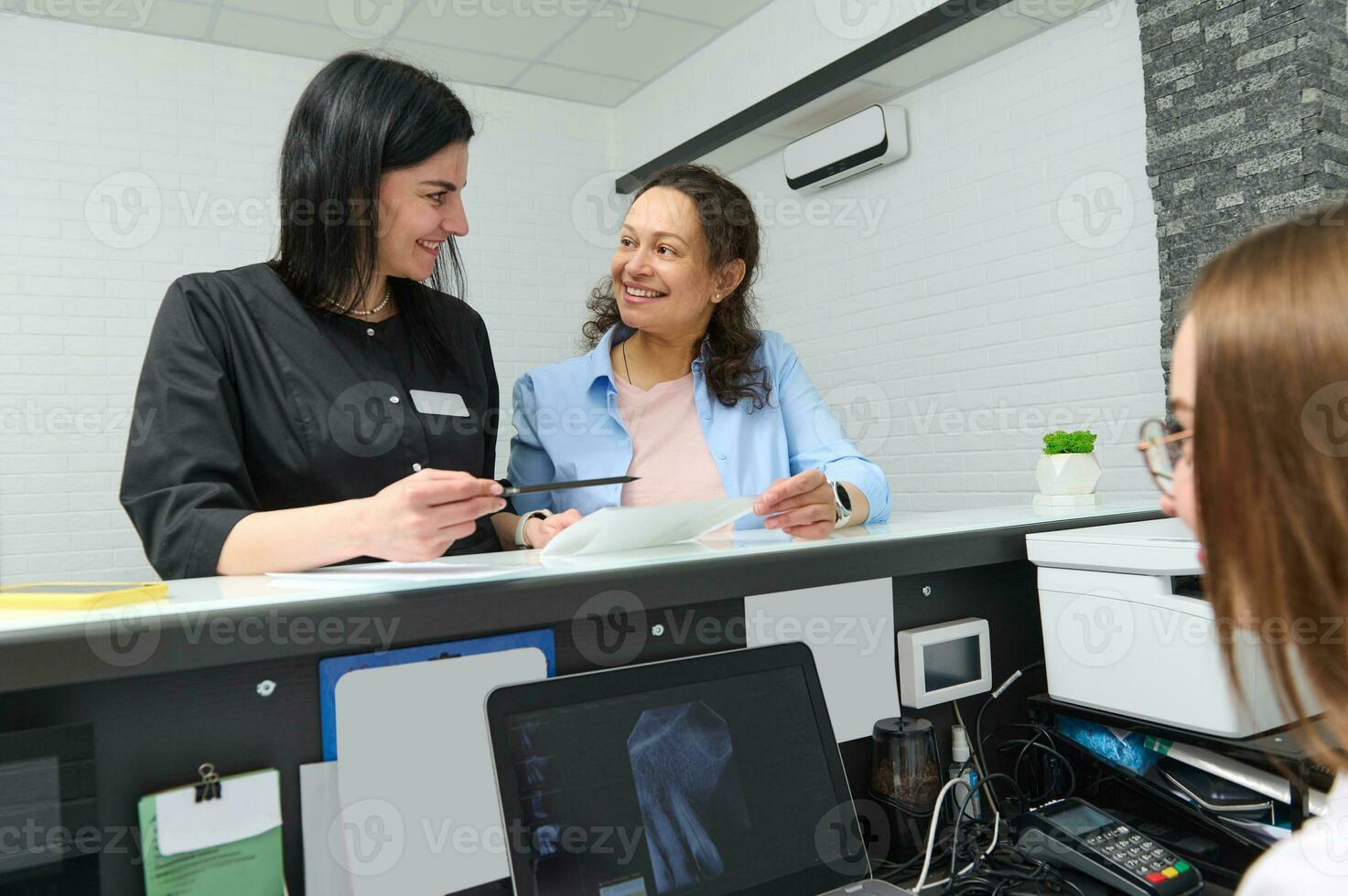 fêmea paciente sorrisos, Perguntando dela médico dentista informações o preenchimento dentro estomatológico forma, em pé de recepção escrivaninha foto