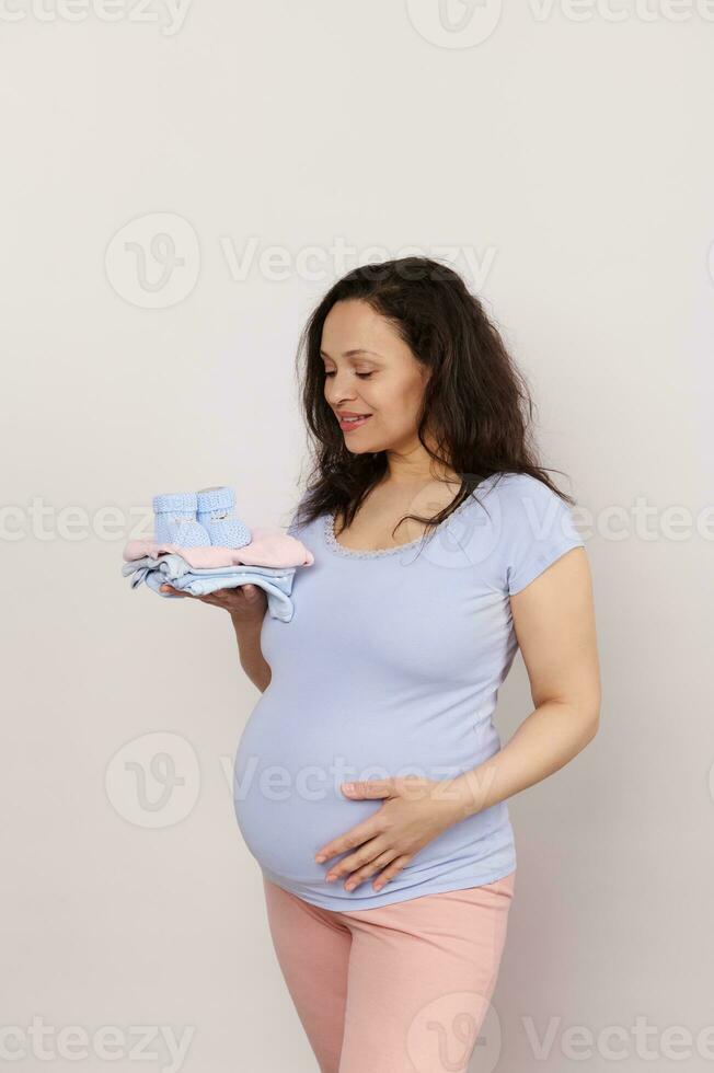 feliz grávida futuro mãe esperando bebê, detém uma pilha do bebê roupas, golpes dela barriga, branco isolado fundo foto