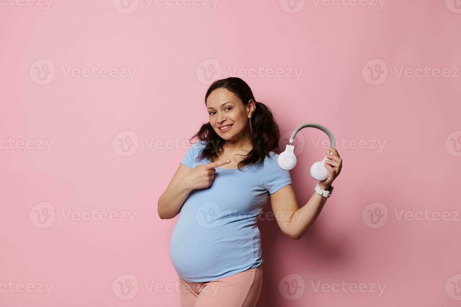 alegre grávida mulher dentro azul camiseta, sorrisos e pontos dedo a parte, de lado sem fio fones de ouvido, isolado em Rosa fundo foto