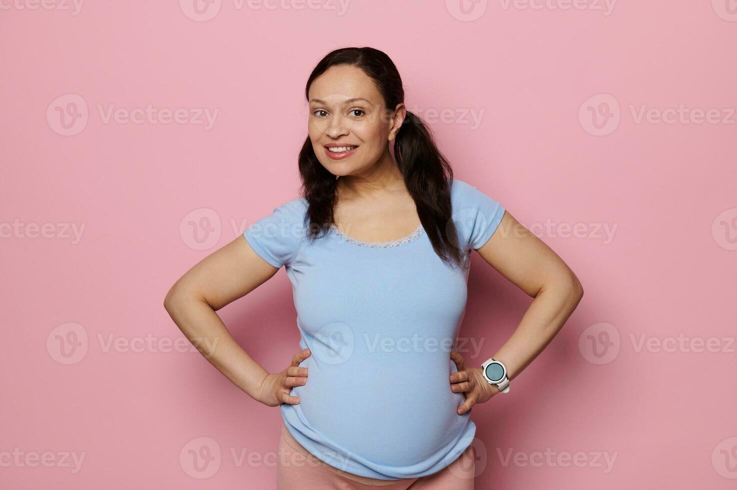 lindo grávida mulher dentro azul camiseta, com grande barriga, sorridente olhando às Câmera, isolado Rosa fundo. gravidez foto