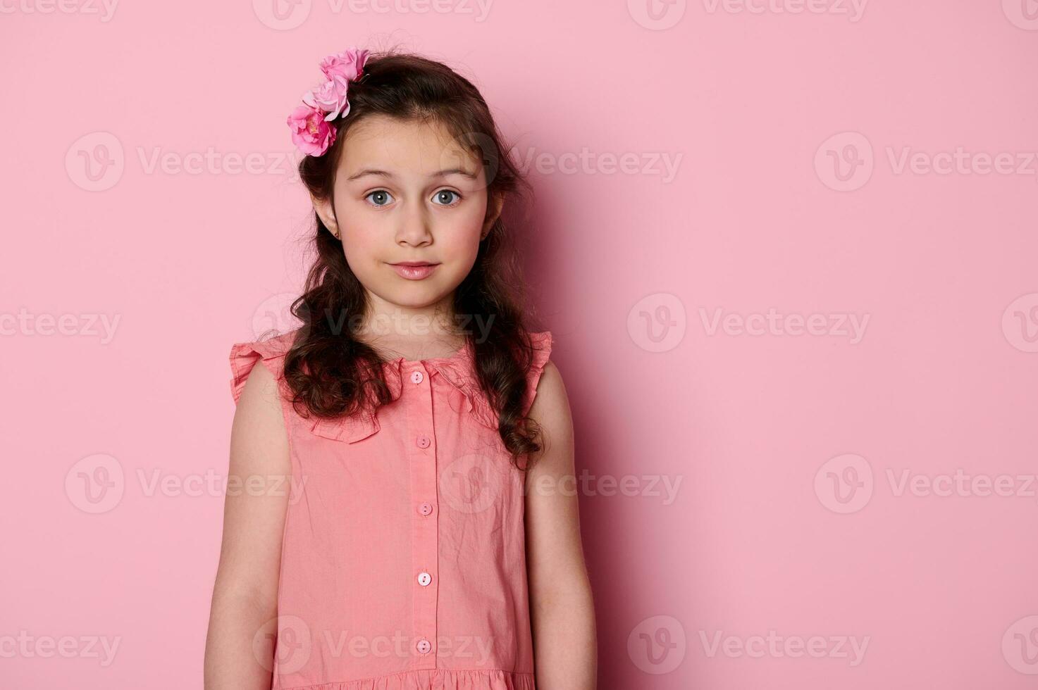 adorável pequeno menina dentro Rosa vestir e rosa flores em cabeça, sorridente olhando às Câmera, isolado em Rosa cor fundo foto