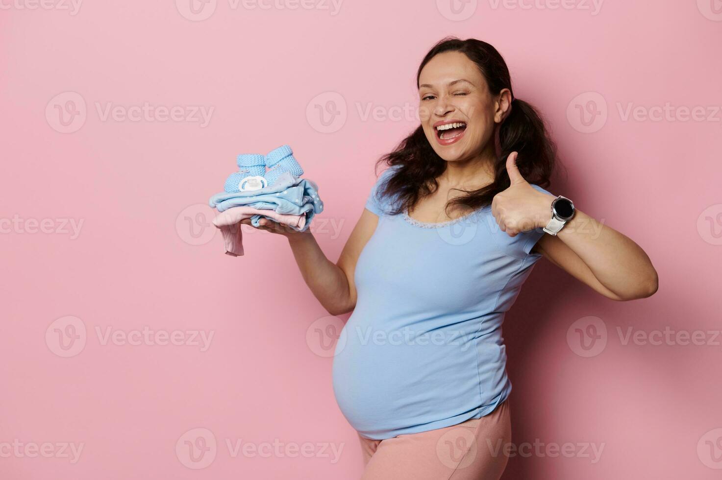expectante mãe detém recém-nascido roupas em dela mão, piscadelas e sorrisos uma cheio de dentes sorrir às Câmera, isolado Rosa fundo foto