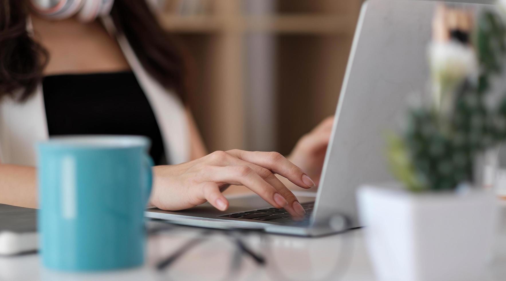 mãos de mulher de negócios digitando, navegando na internet no teclado do computador portátil com smartphone móvel, fones de ouvido, uma xícara de café na mesa do escritório de madeira no escritório em casa. aluna aprendendo online foto