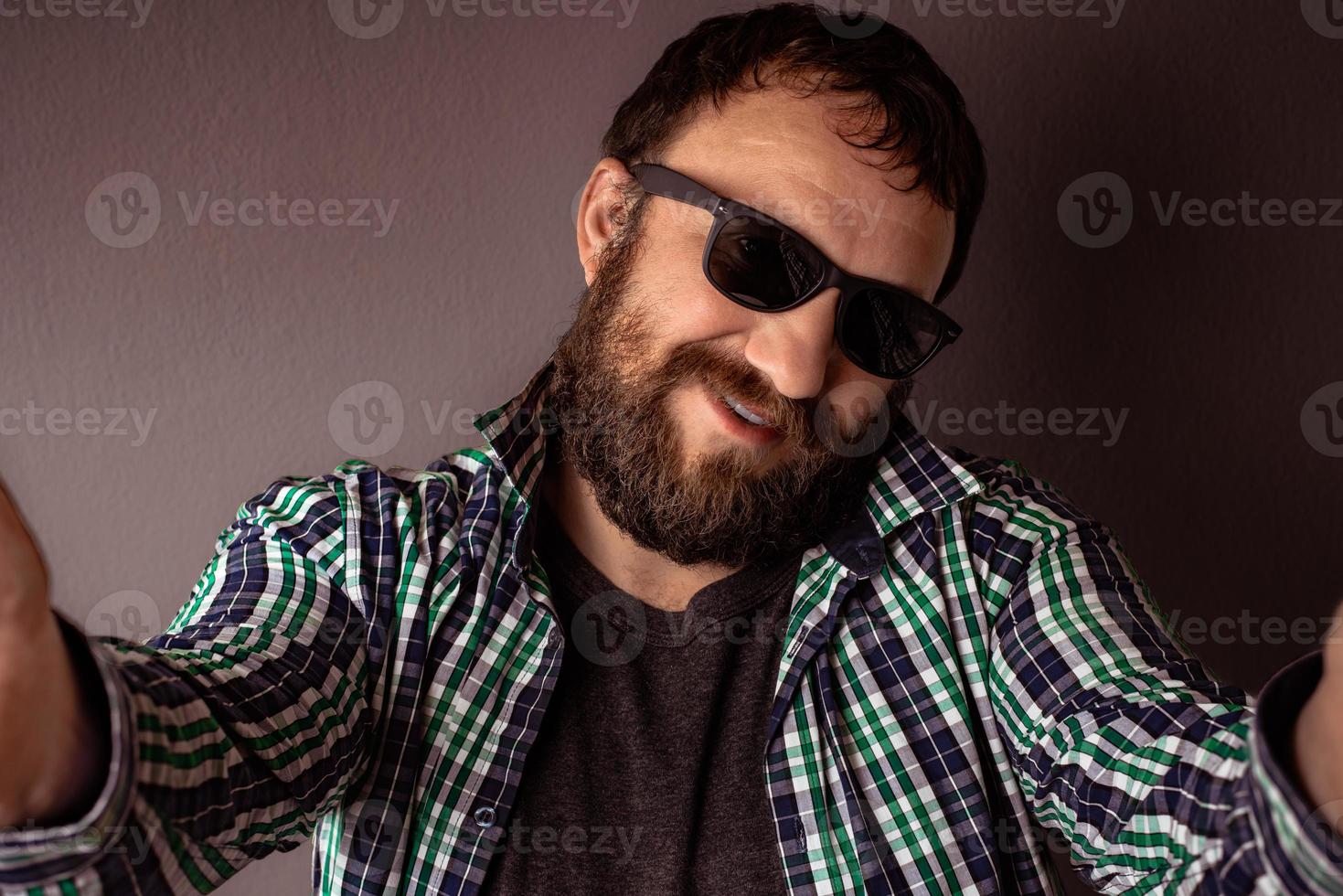 hipster bonito barbudo homem com chapéu, óculos escuros e camisa tirando uma selfie foto