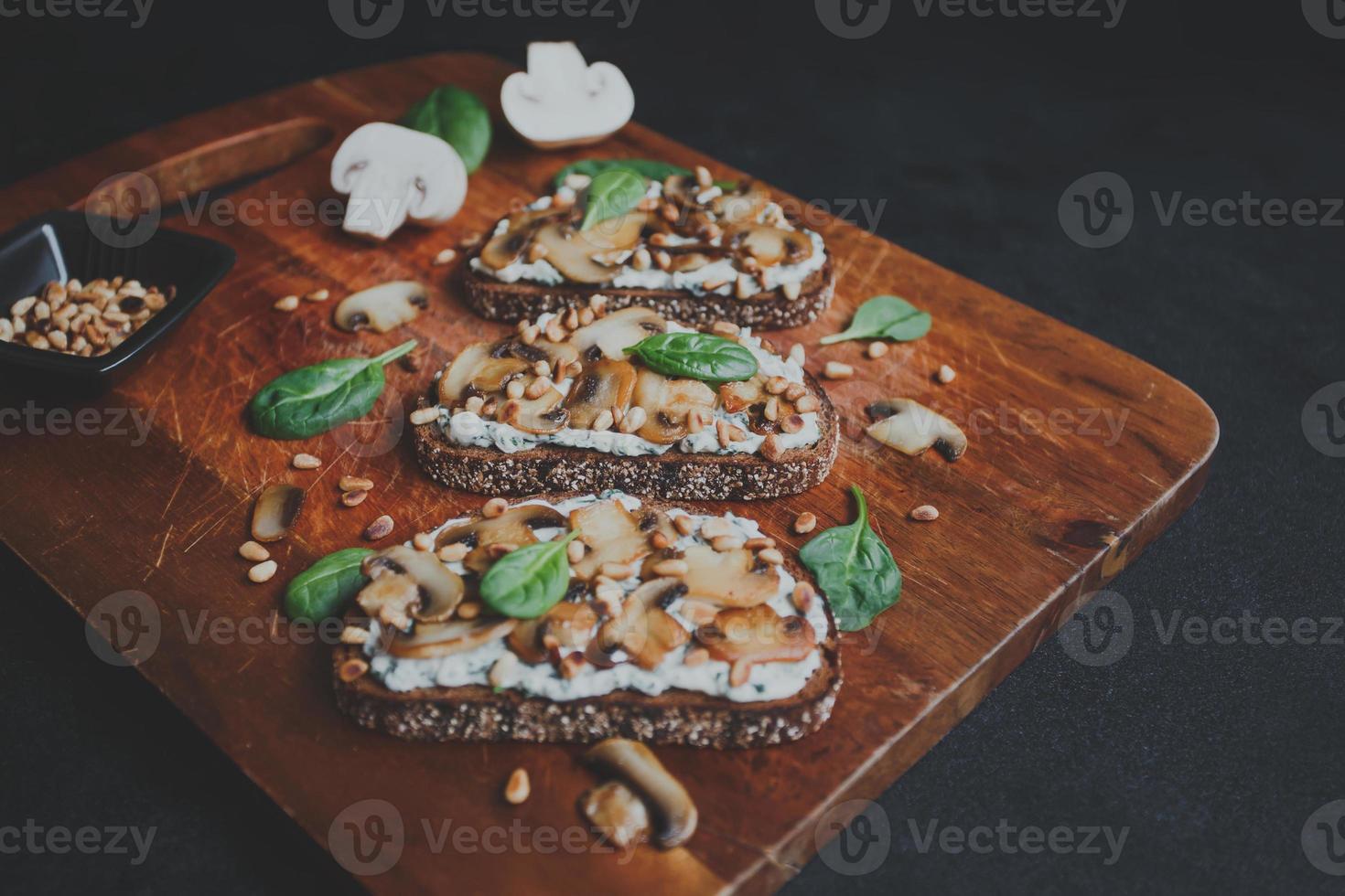 saborosa bruschetta fresca com cogumelos, espinafre, alho, cream cheese e pinhões, em uma placa de madeira, sobre um fundo escuro. foto