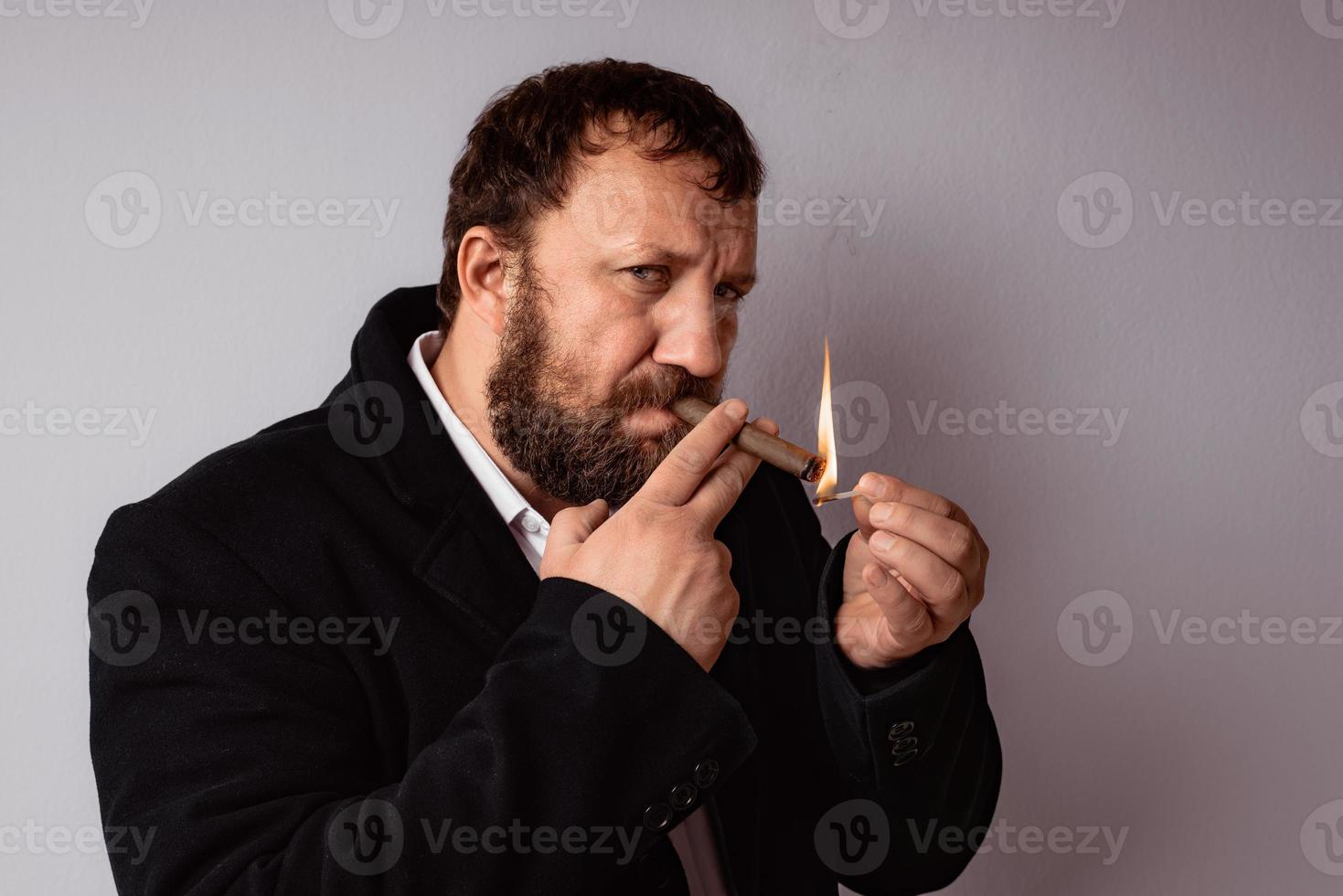 homem barbudo com casaco e camisa modernos acendendo o charuto foto