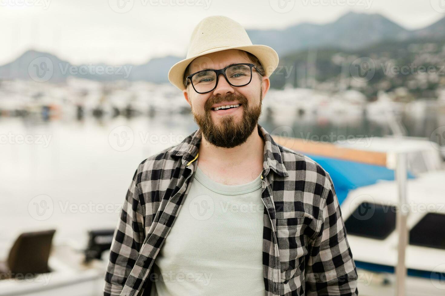 bonito homem vestindo chapéu e óculos perto marina com iates. retrato do rindo homem com mar porta fundo foto