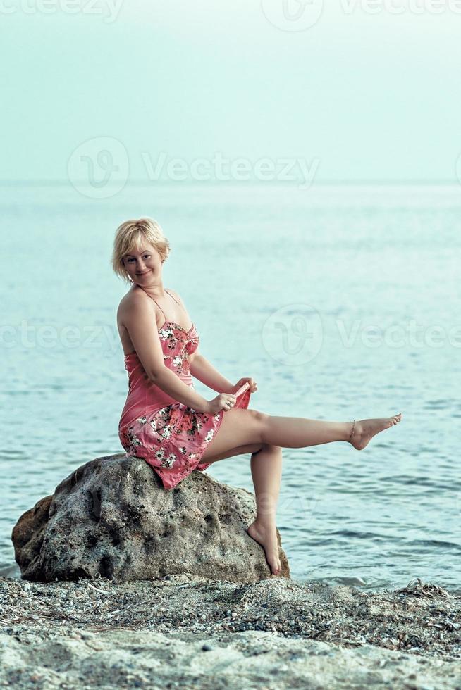 mulher loira feliz em um vestido florido avermelhado. mulher sentada e posar na grande pedra no litoral. foto