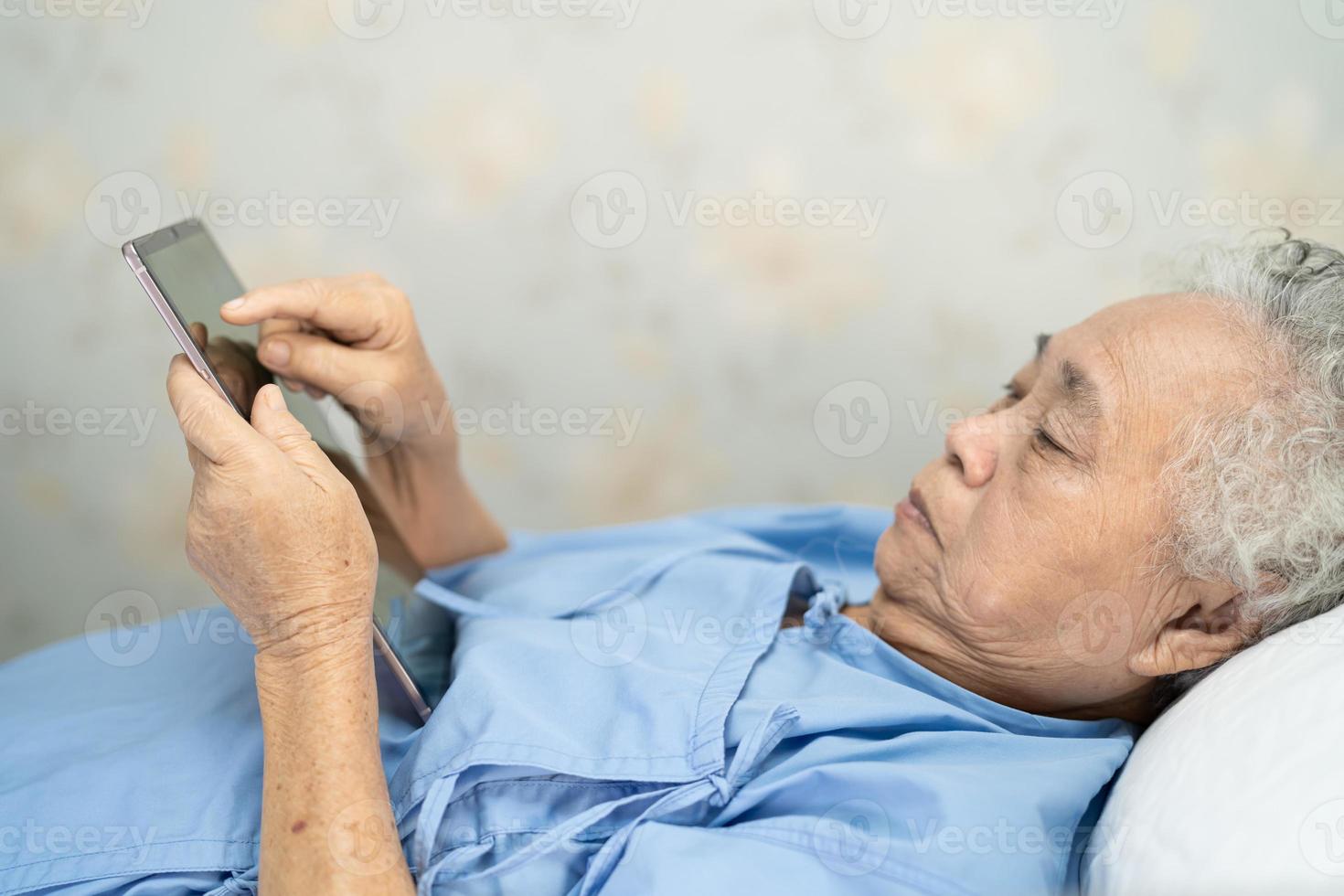 paciente idoso asiático ou idosa segurando um tablet digital nas mãos e lendo e-mails enquanto está sentado na cama na enfermaria do hospital de enfermagem, conceito médico forte e saudável foto