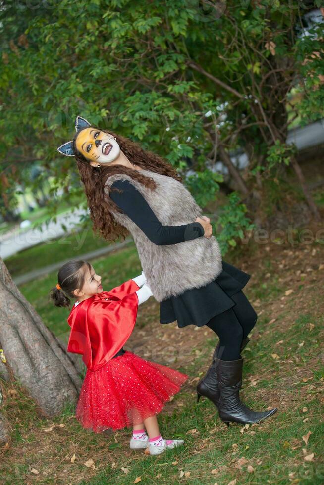 lindo menina em pequeno vermelho equitação de capuz traje jogando com dela mãe. real família tendo Diversão enquanto usando fantasias do a pequeno vermelho equitação de capuz conto dentro dia das Bruxas. foto