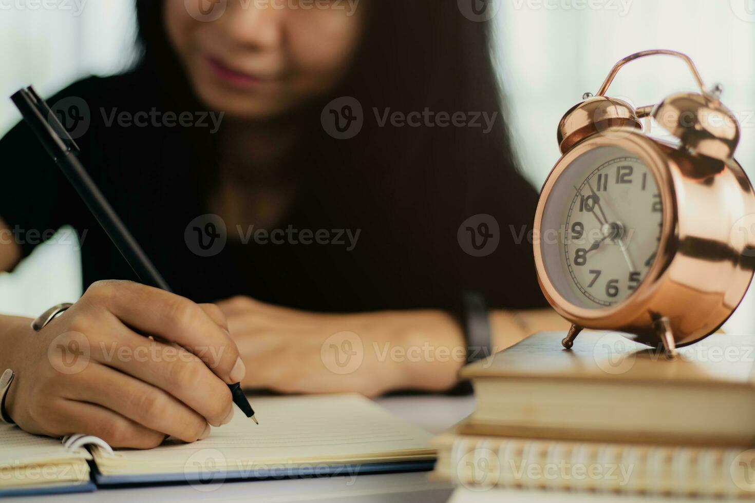 ásia mulher escrevendo em uma caderno com Sino alarme relógio em livros foto