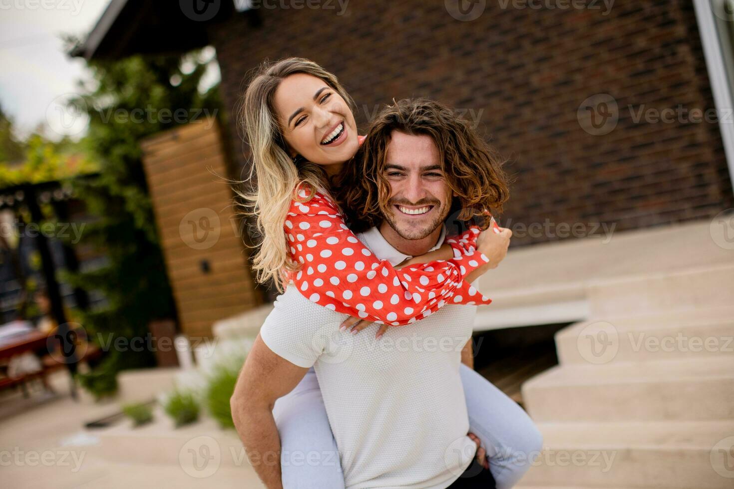 bonito grandes cabelo homem carregando a jovem mulher em dele costas dentro frente do tijolo casa foto