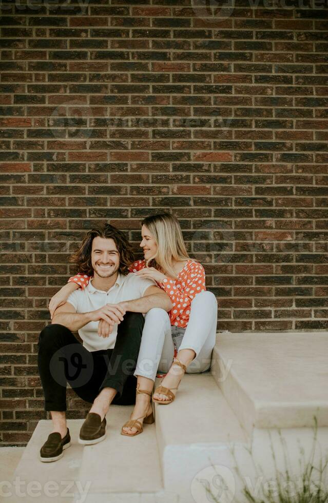 sorridente jovem casal dentro amor sentado dentro frente do casa tijolo parede foto