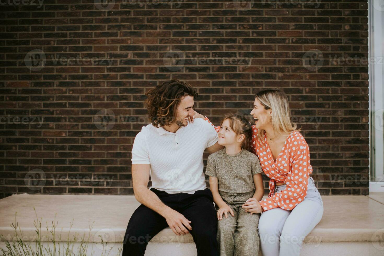 família com uma mãe, pai e filha sentado lado de fora em a passos do uma frente varanda do uma tijolo casa foto