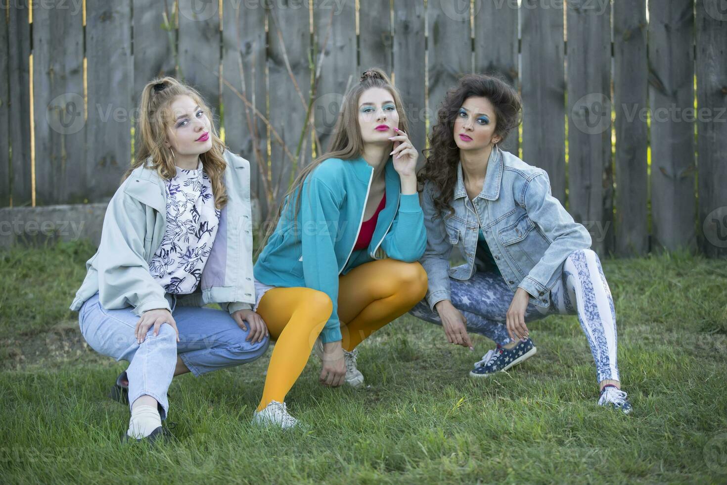 engraçado meninas dentro a estilo do a anos 90 dentro brilhante roupas estão sentado perto uma de madeira cerca. foto