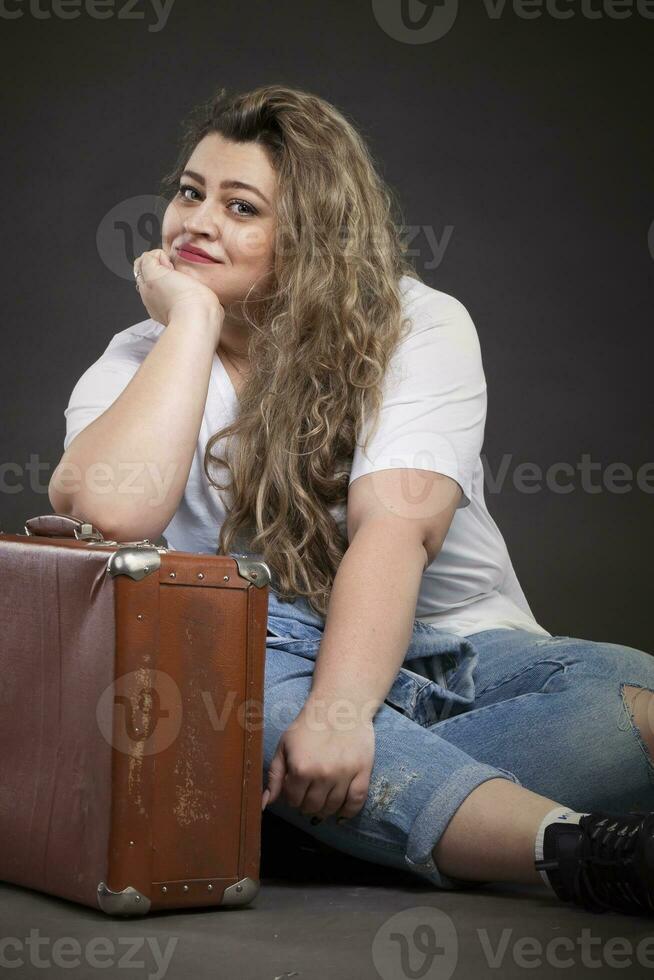 lindo gordo mulher dentro jeans roupas com a velho retro mala de viagem em uma cinzento fundo. mais Tamanho modelo garota. foto