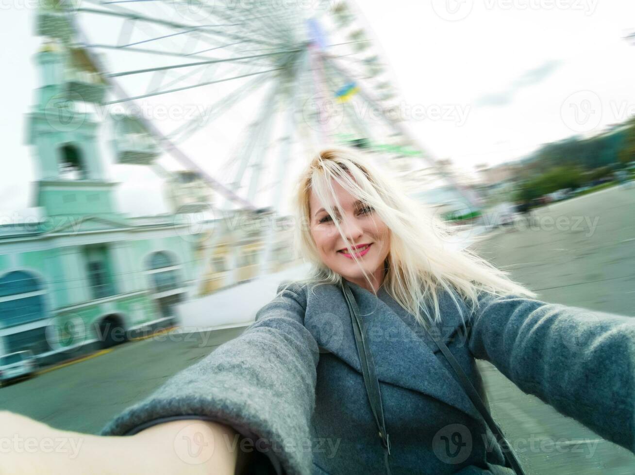 atraente Loiras menina dentro casual roupas, vestindo uma casaco, leva selfie, olhando para dentro a Câmera. à moda menina leva selfie, fundo do a ferris roda foto