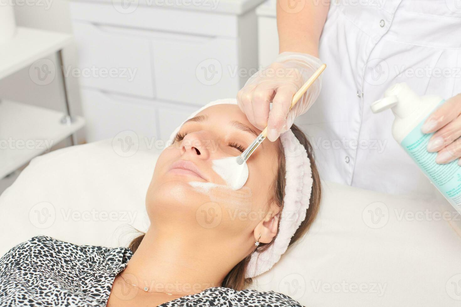 cosmetologista aplicando máscara de creme cosmético no rosto feminino para procedimento de rejuvenescimento no salão de beleza foto