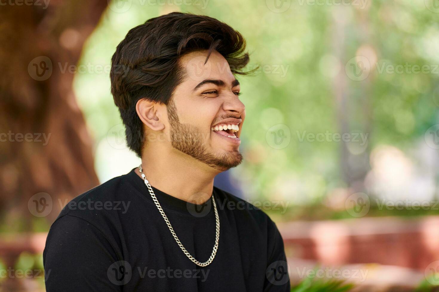 sorridente jovem indiano homem retrato dentro Preto t camisa e prata pescoço cadeia ao ar livre verde público parque foto