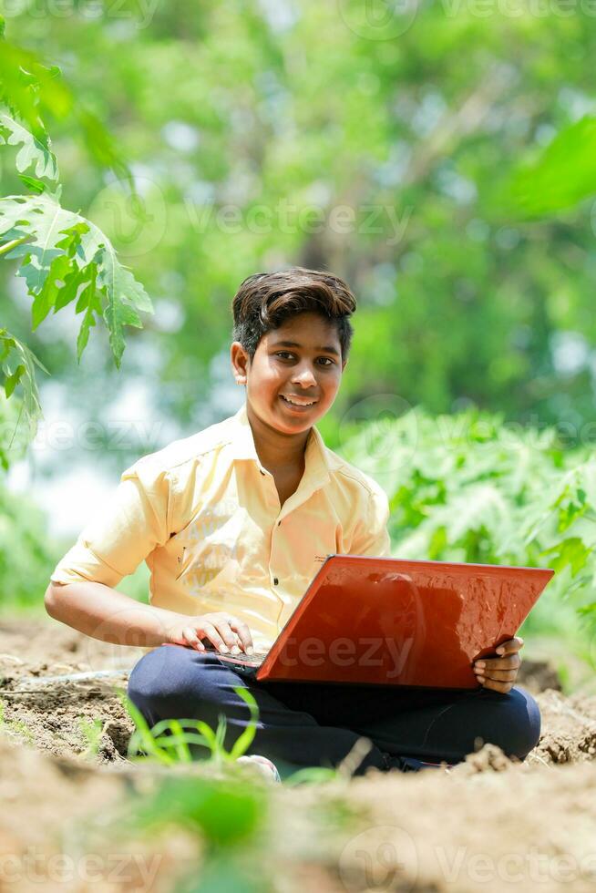 indiano Garoto estudando dentro fazenda, segurando computador portátil dentro mão , pobre indiano crianças foto