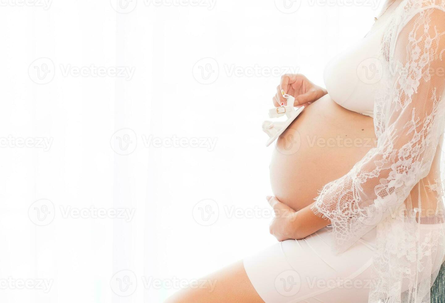 fechar-se grávida barriga com fofa minúsculo bebê sapato. conceito do gravidez, ginecologia, preparação. lindo grávida mulher dentro camisa dentro branco fundo. mulher segurando grande grávida estômago. foto