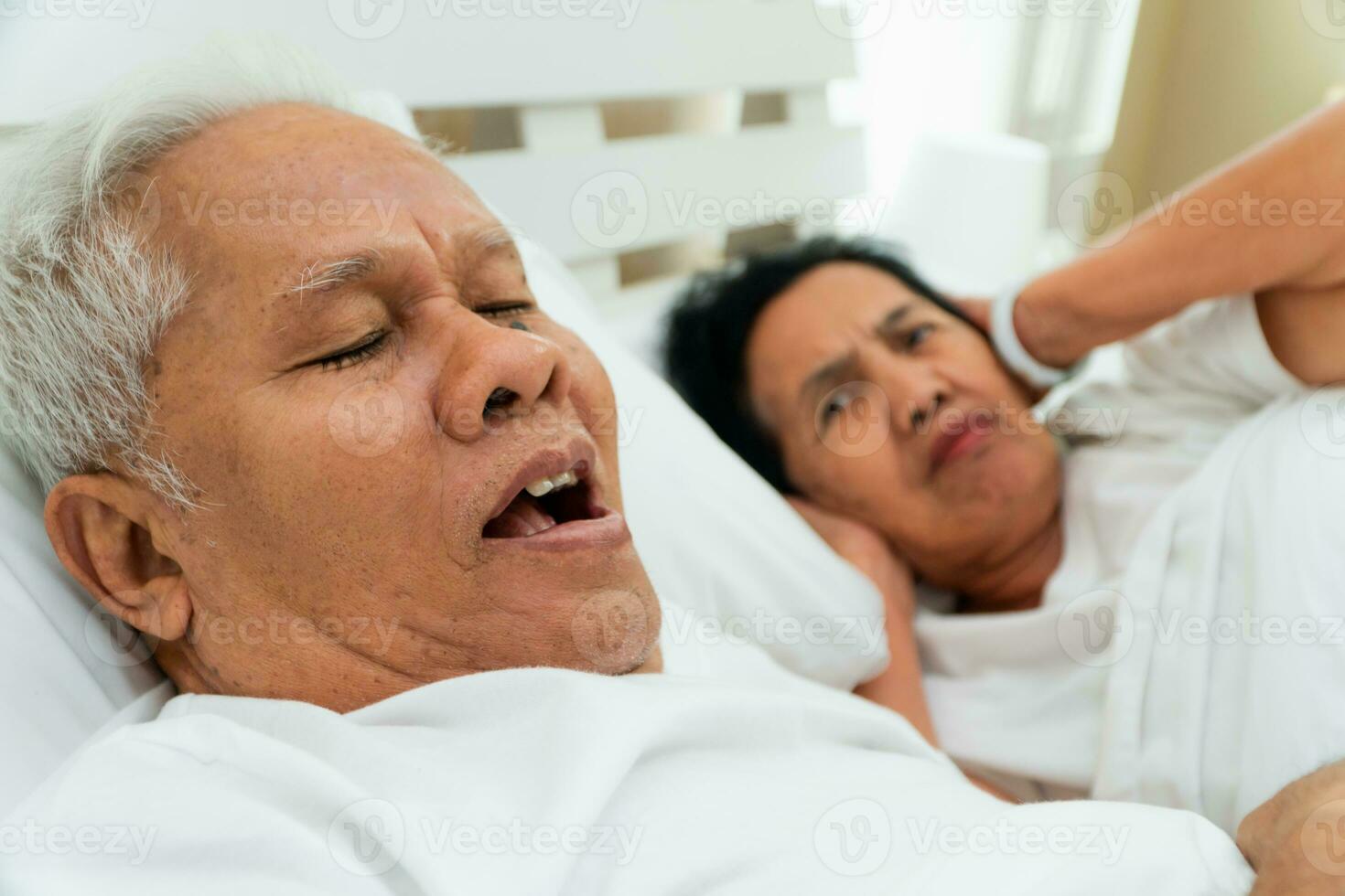 Mais velho ásia mulher deitado aberto dela olhos e cobrir orelhas com dela mãos dentro cama ao lado dela marido quem ronca e faz barulho, conjugal problemas. foto