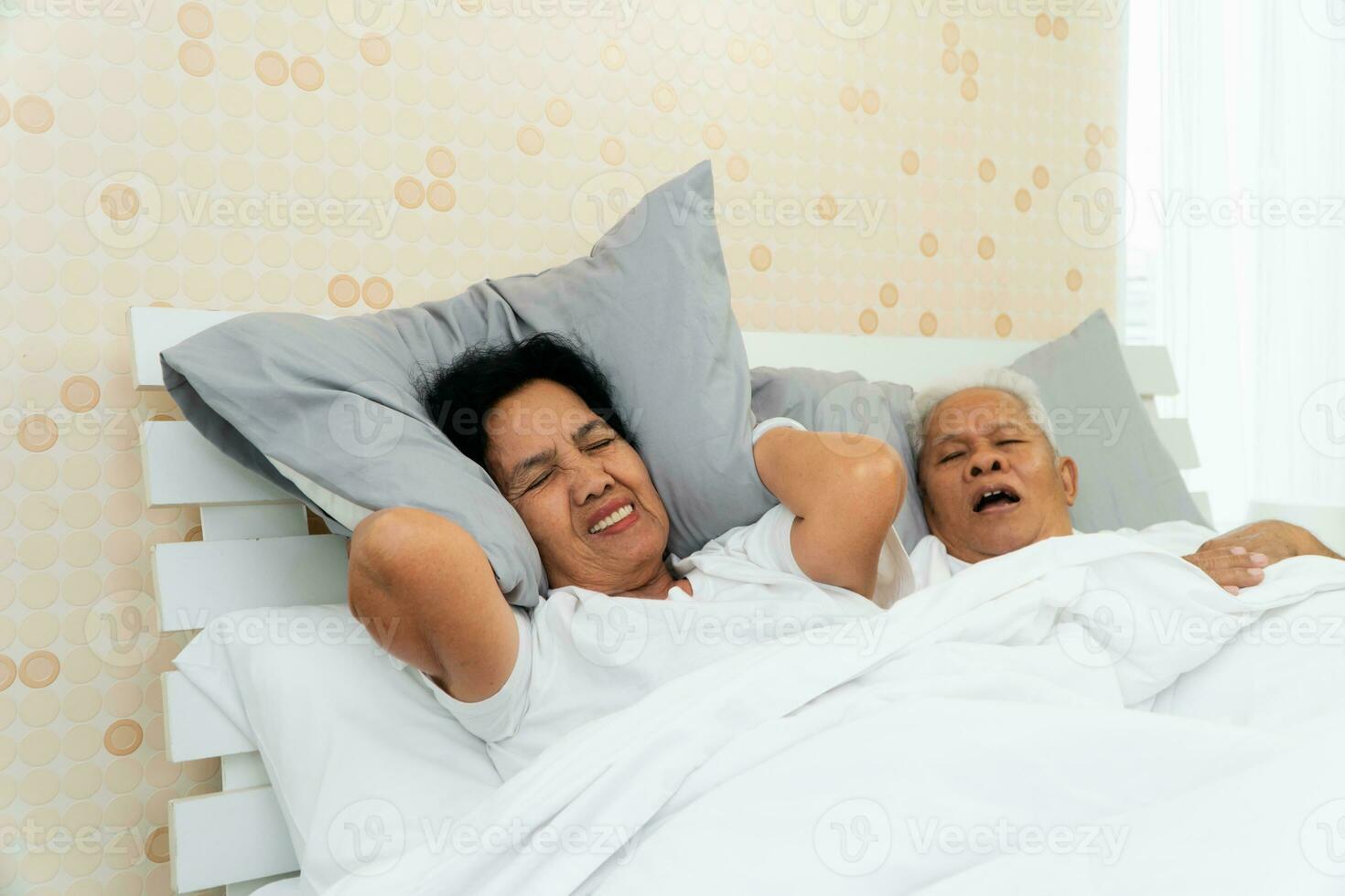 Mais velho ásia mulher deitado aberto dela olhos e cobrir orelhas com dela travesseiro dentro cama ao lado dela marido quem ronca e faz barulho, conjugal problemas. foto