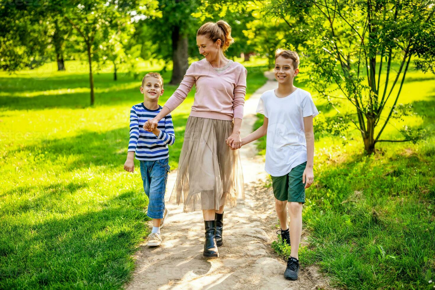 feliz mãe é caminhando com dela filhos dentro parque. elas estão segurando mãos e desfrutando verão dia. foto