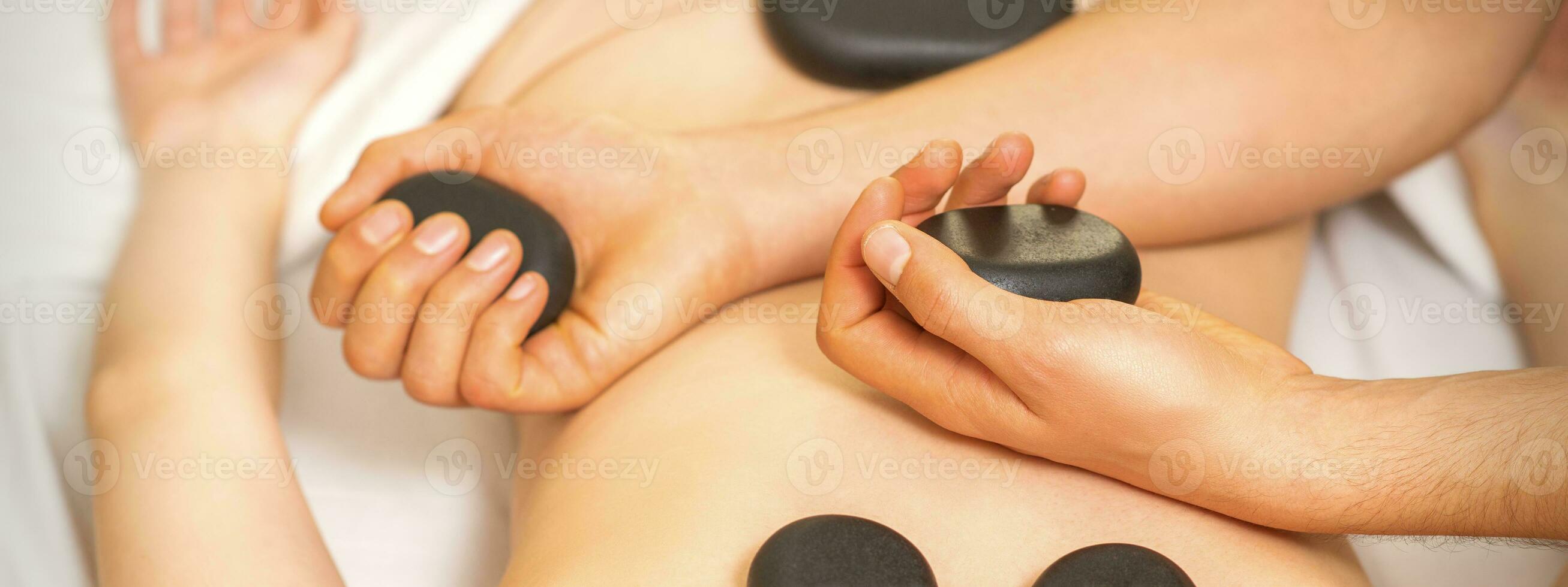 quente pedra massagem em a fêmea costas com mãos do massagista segurando Preto massagem pedras dentro spa salão. foto