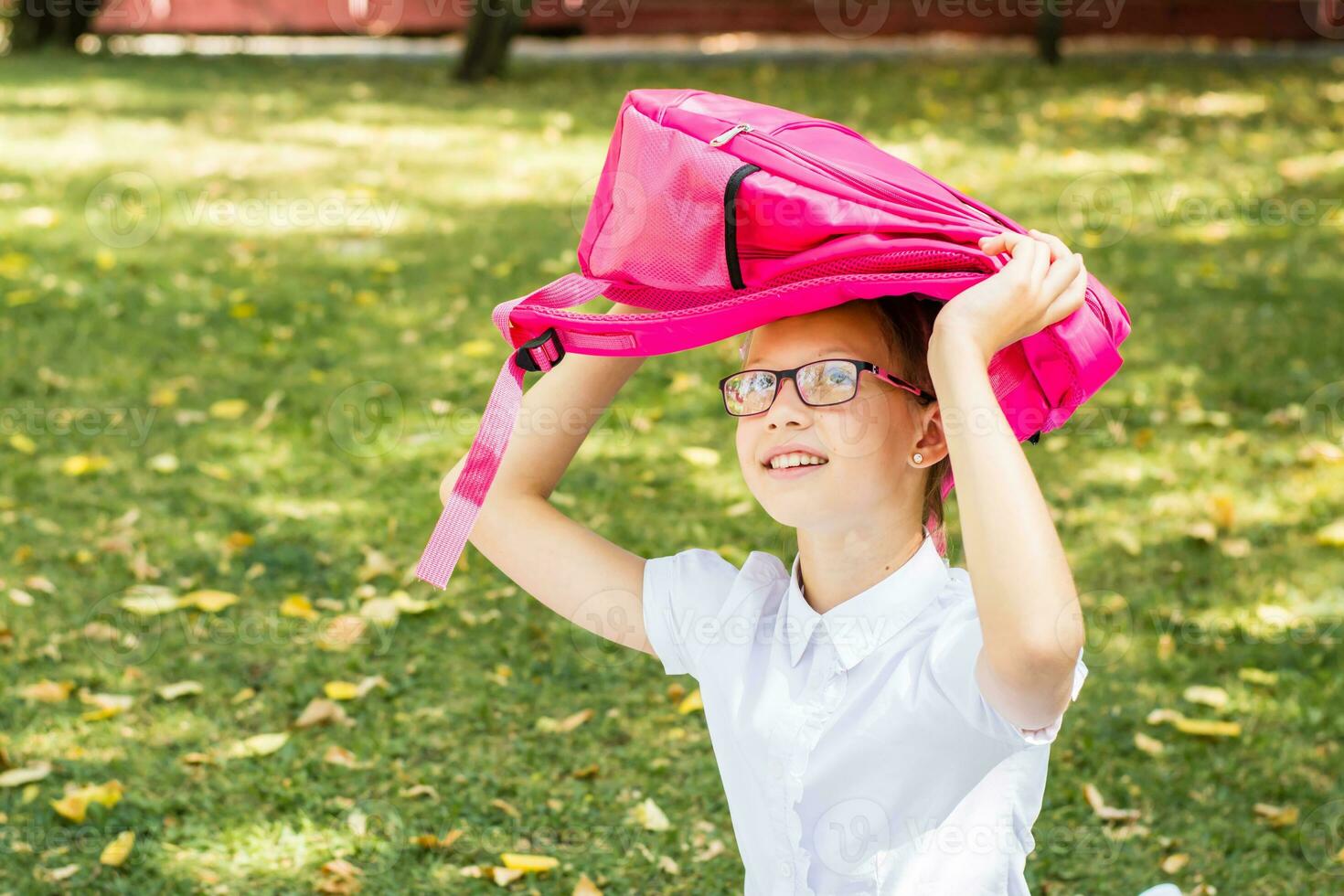 uma fofa estudante com óculos sorrisos e detém uma mochila acima dela cabeça dentro uma ensolarado outono parque. costas para escola conceito. cópia de espaço foto