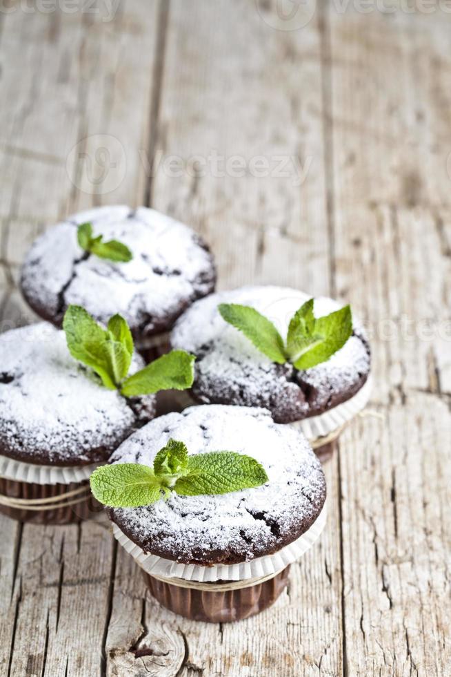 muffins escuros de chocolate frescos com açúcar em pó e folha de hortelã na mesa de madeira rústica. foto