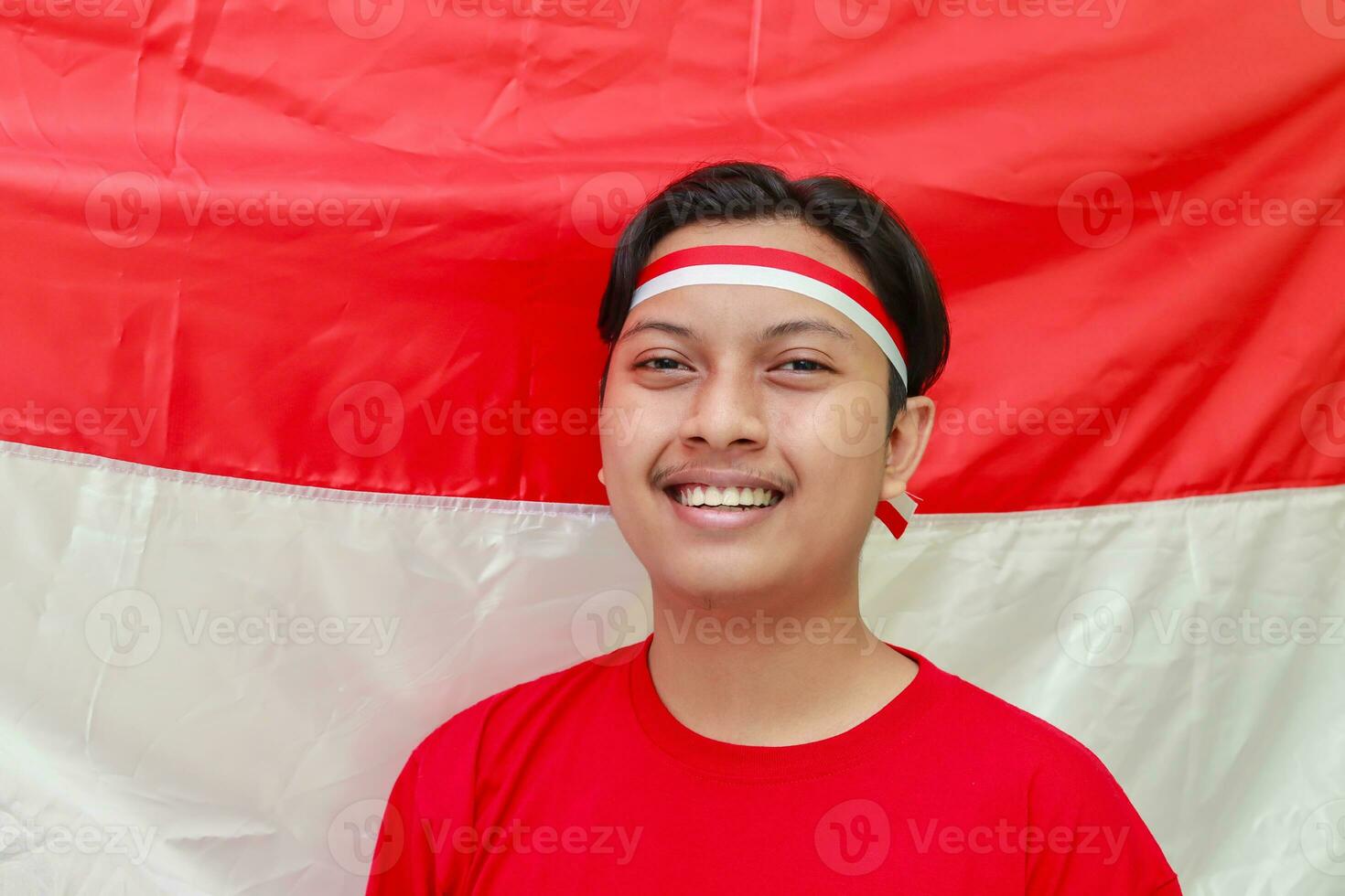 retrato do atraente ásia homem vestindo camiseta com vermelho e branco fita em cabeça, em pé dentro frente do indonésio bandeira e olhando às Câmera foto