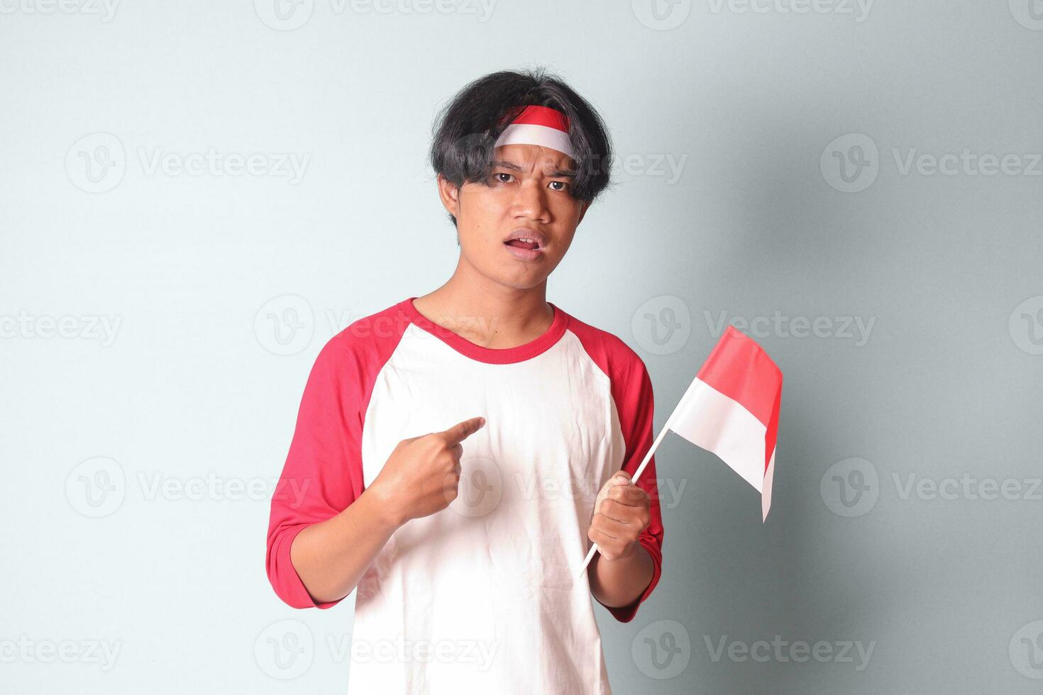 retrato do atraente ásia homem dentro camiseta com vermelho e branco fita em cabeça, mostrando produtos e poiting longe enquanto segurando indonésio bandeira. isolado imagem em cinzento fundo foto