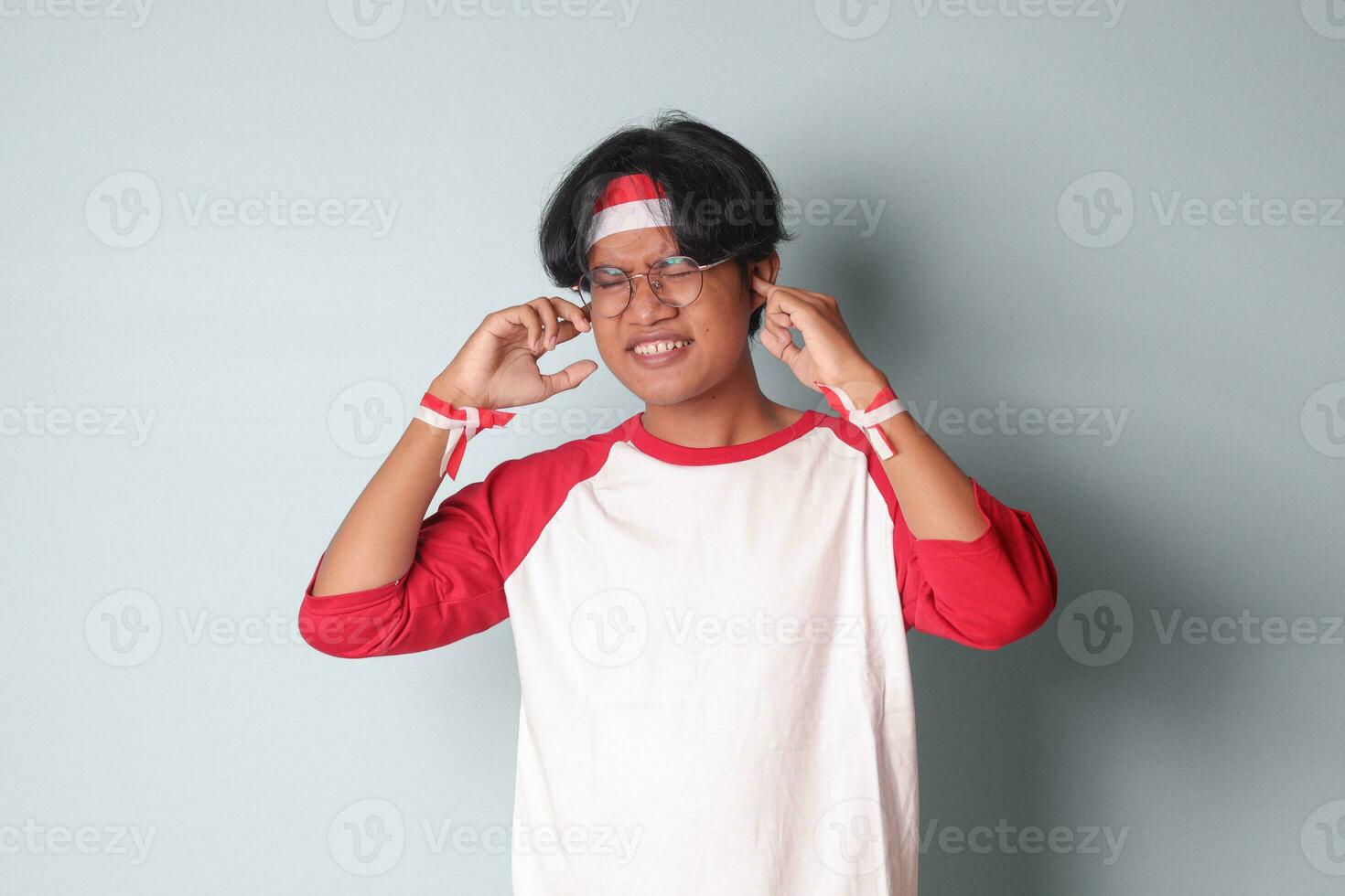 retrato do atraente ásia homem dentro camiseta com vermelho e branco fita em cabeça, tendo uma enxaqueca, tocante dele têmpora. dor de cabeça conceito. isolado imagem em cinzento fundo foto