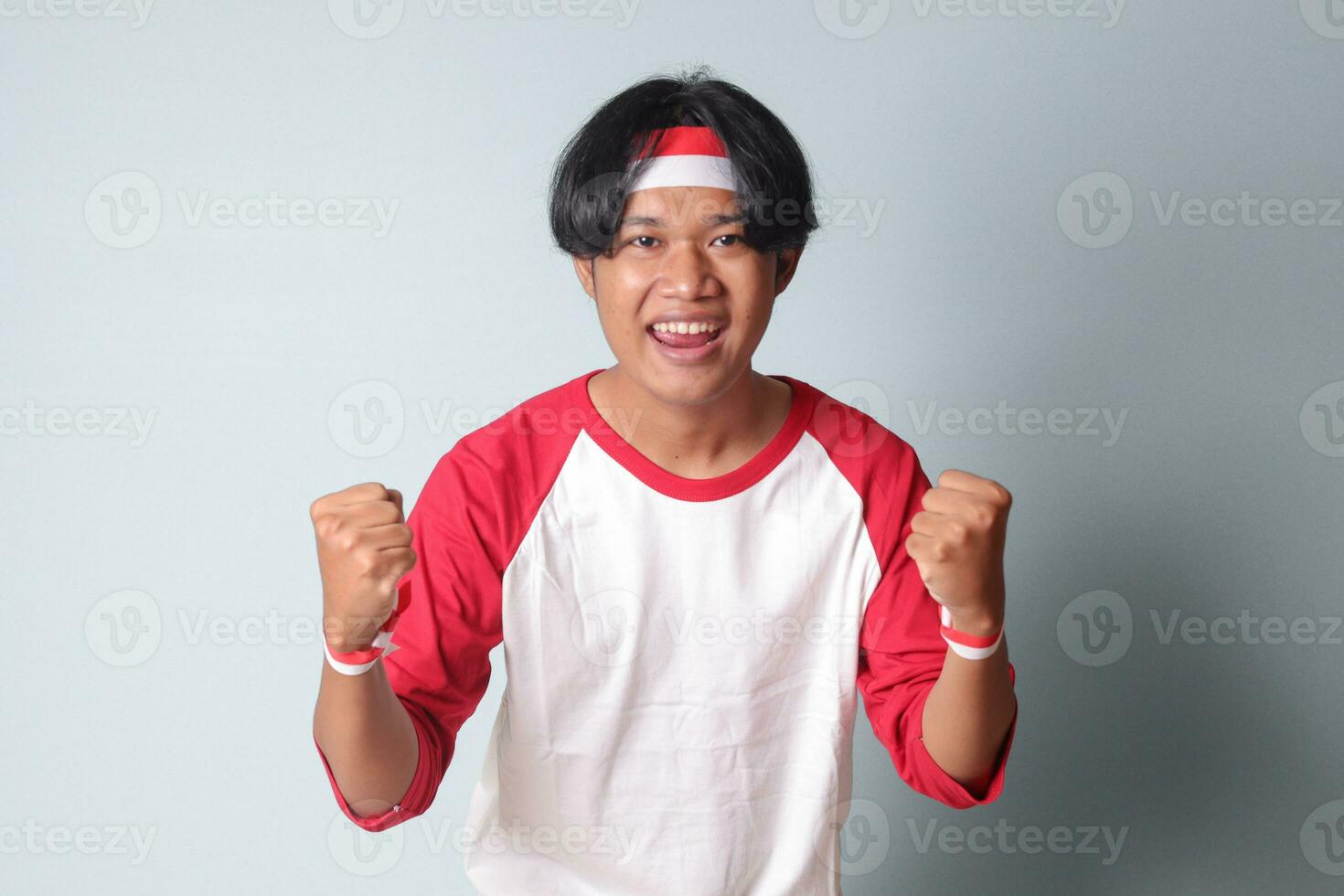 retrato do atraente ásia homem dentro camiseta com vermelho e branco fita em cabeça, levantando dele punho, a comemorar da indonésia independência dia. isolado imagem em cinzento fundo foto