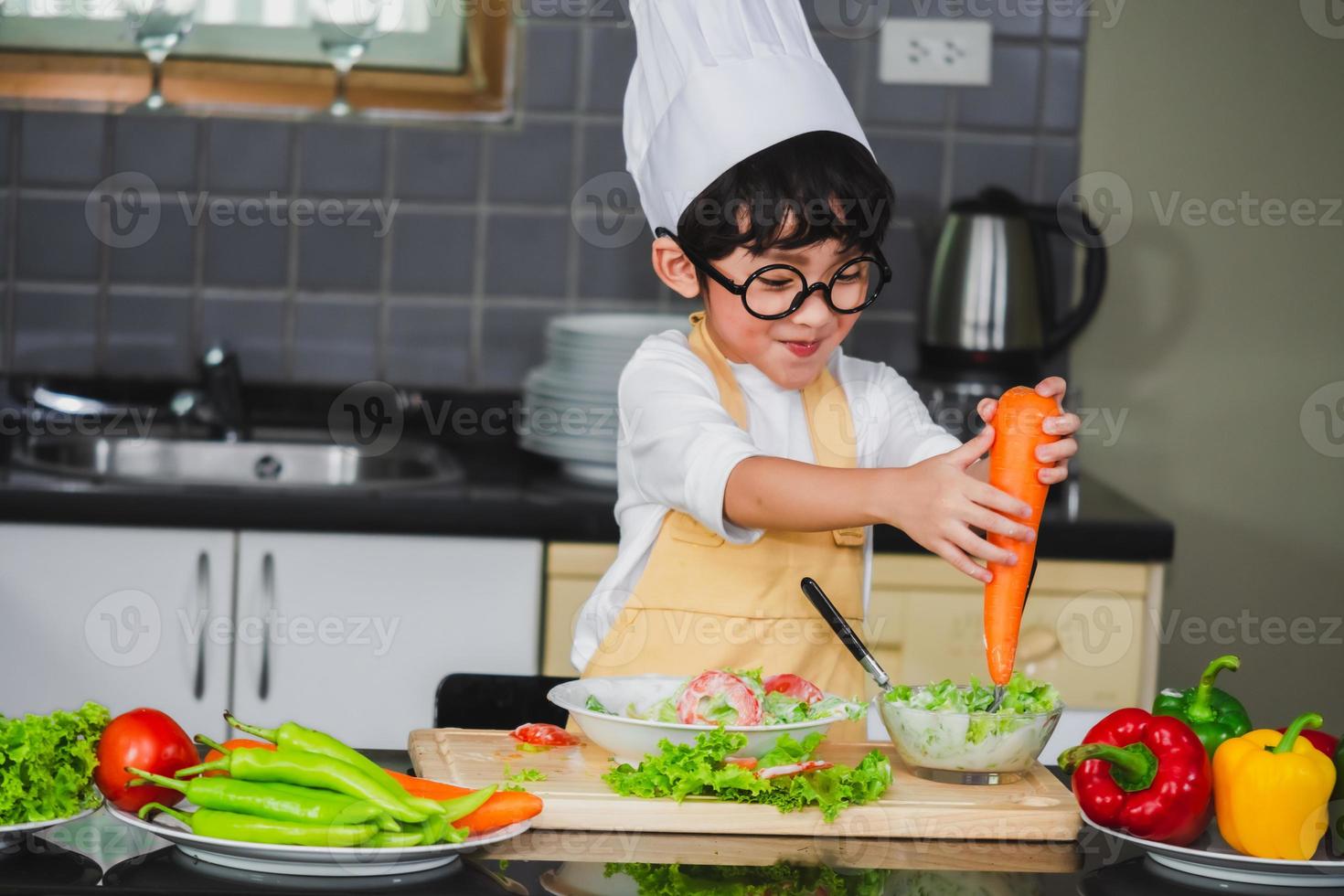 filho de menino asiático cozinhando salada de comida holdind colher de madeira com vegetais segurando tomates e cenouras, pimentões no prato para família feliz cozinhar comida prazer estilo de vida foto